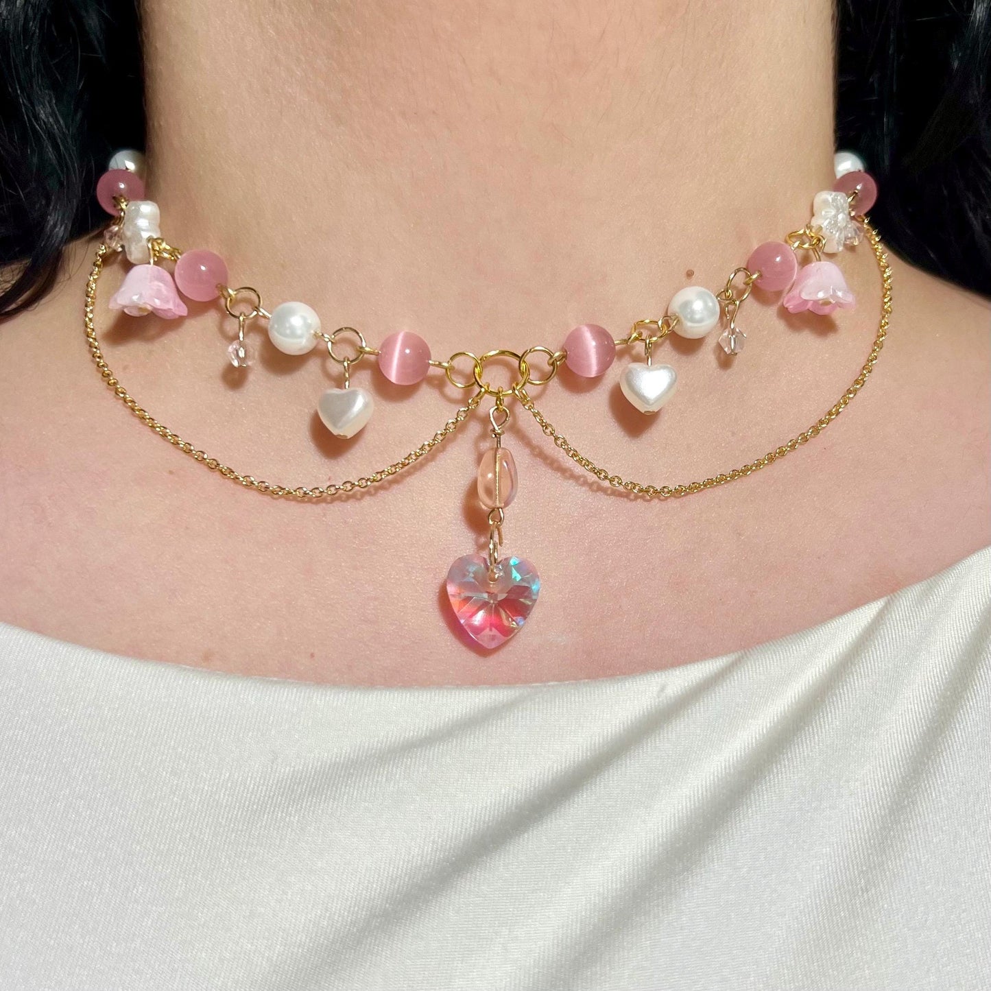 Close-up of gold-plated choker necklace featuring a sparkling iridescent heart pendant, surrounded by pink and pearl charms with floral details.