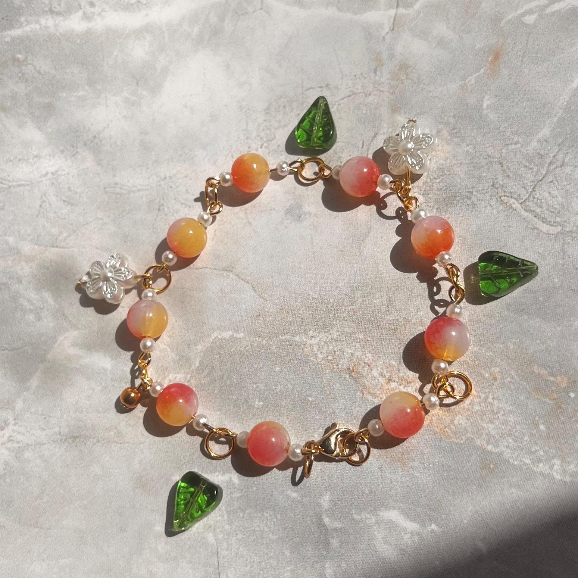a bracelet made of pink and orange beads with white pearl accents. The bracelet has a gold clasp and is decorated with green and white charms in the shape of leaves and flowers.
