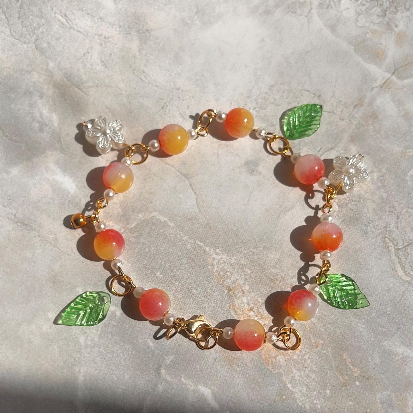 A peach-colored bracelet with gold chain links, white pearl beads, and green leaf and white flower charms.
