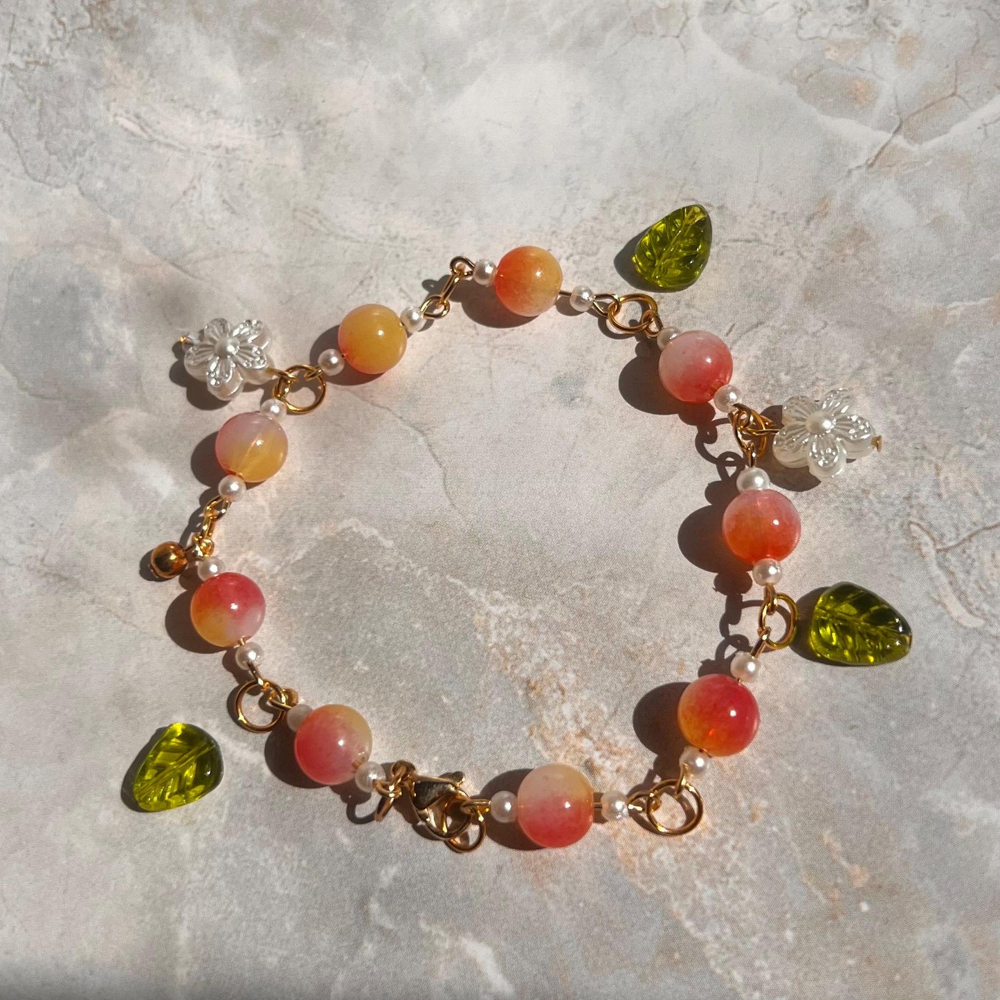 A gold chain bracelet with pink and orange beads, white flower charms, and green leaf charms.