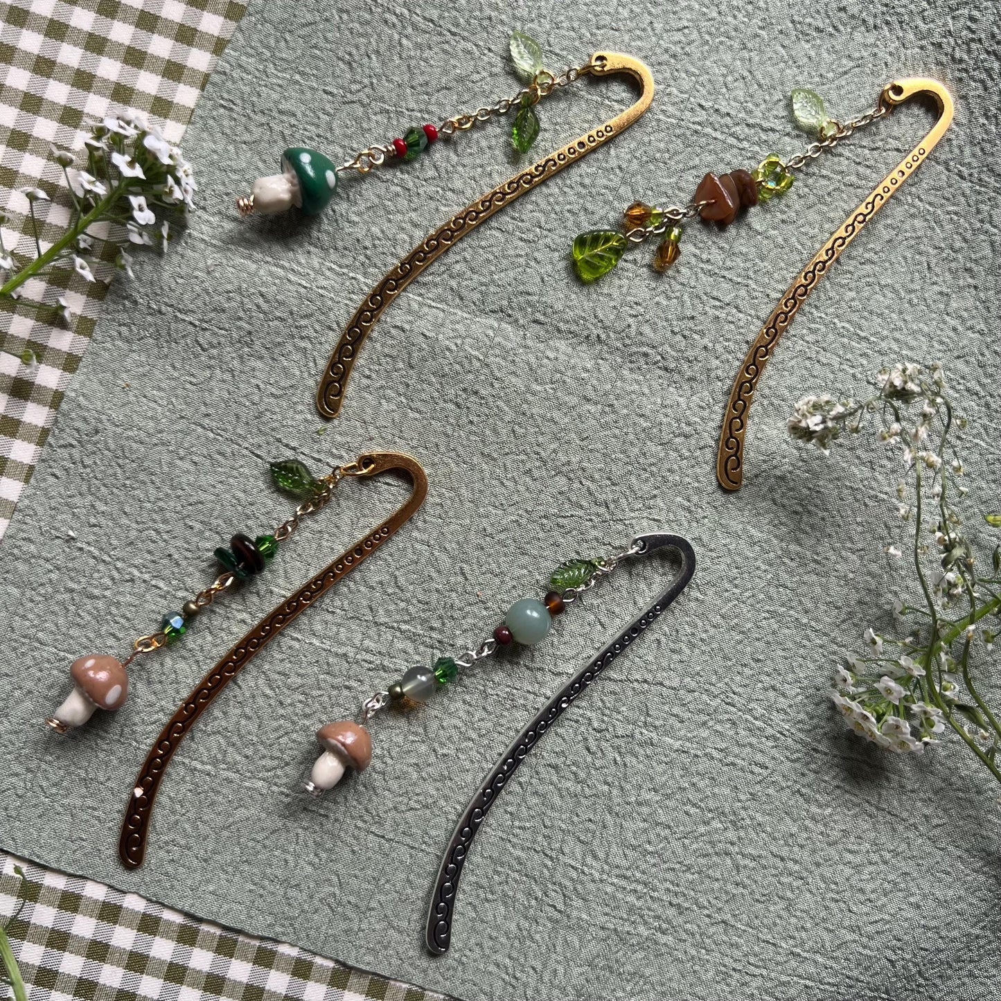 Five handmade metal bookmarks with gold and bronze finishes, decorated with green, brown, and red-toned beaded charms. Each bookmark features forest-themed details like glass leaves, gemstone chips, and small polymer clay mushrooms. Displayed on a sage green textured fabric with small white flowers