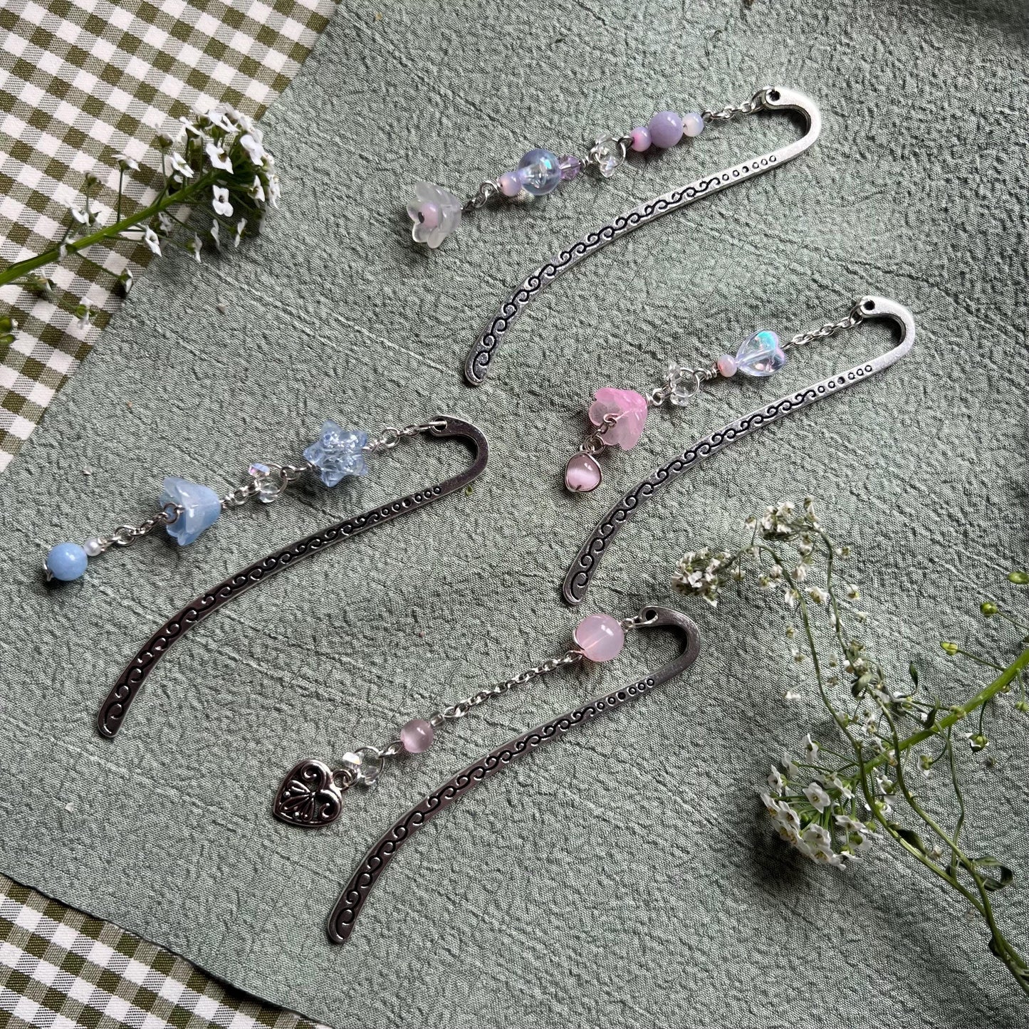 five silver-toned handmade metal bookmarks with soft pastel beaded charms in shades of pink, blue, and purple. Designs include glass flowers, heart charms, and delicate accent beads. Set on a sage green textured background with white wildflowers, evoking a dreamy, fairycore aesthetic