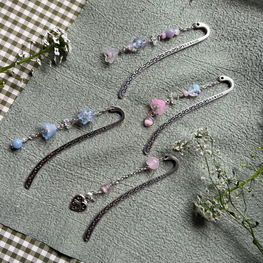 five silver-toned handmade metal bookmarks with soft pastel beaded charms in shades of pink, blue, and purple. Designs include glass flowers, heart charms, and delicate accent beads. Set on a sage green textured background with white wildflowers, evoking a dreamy, fairycore aesthetic